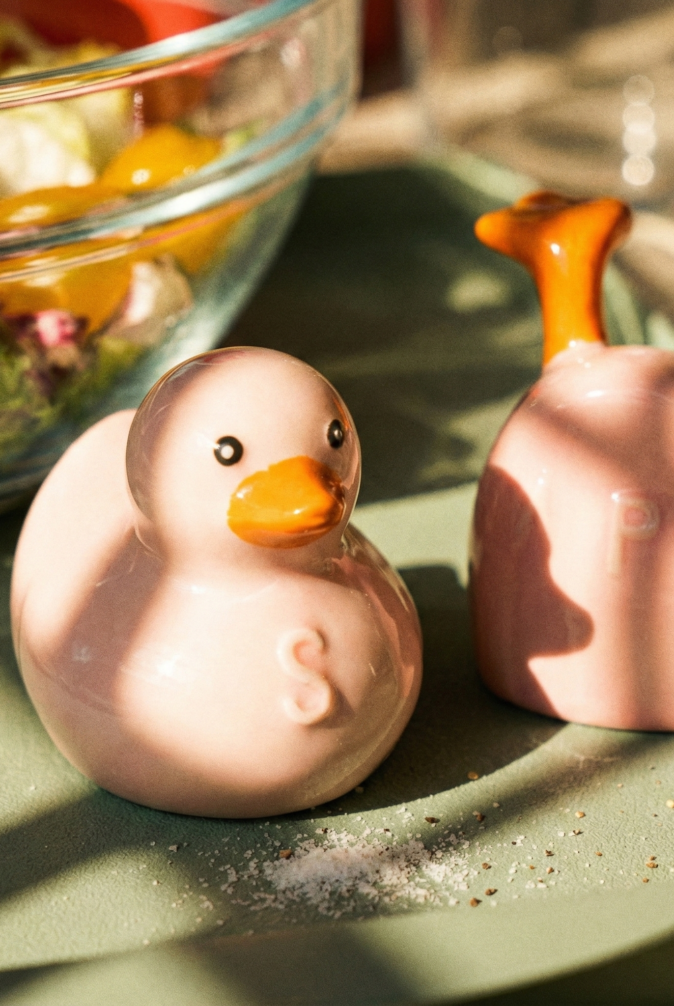 Two ceramic duck-shaped salt and pepper shakers on a green surface with a salad in the background.