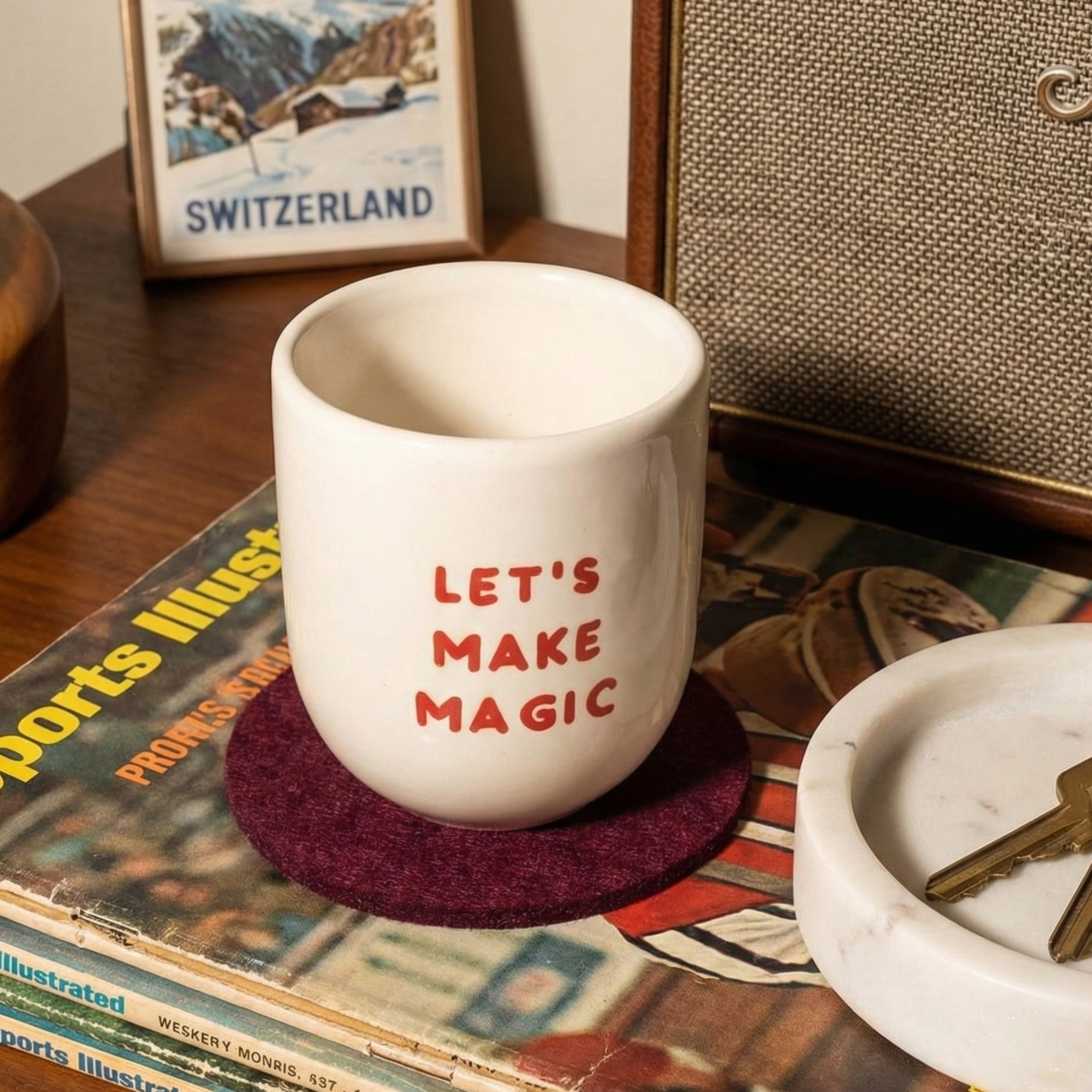 White mug with 'Let's Make Magic' text on a wooden surface with a magazine and key.