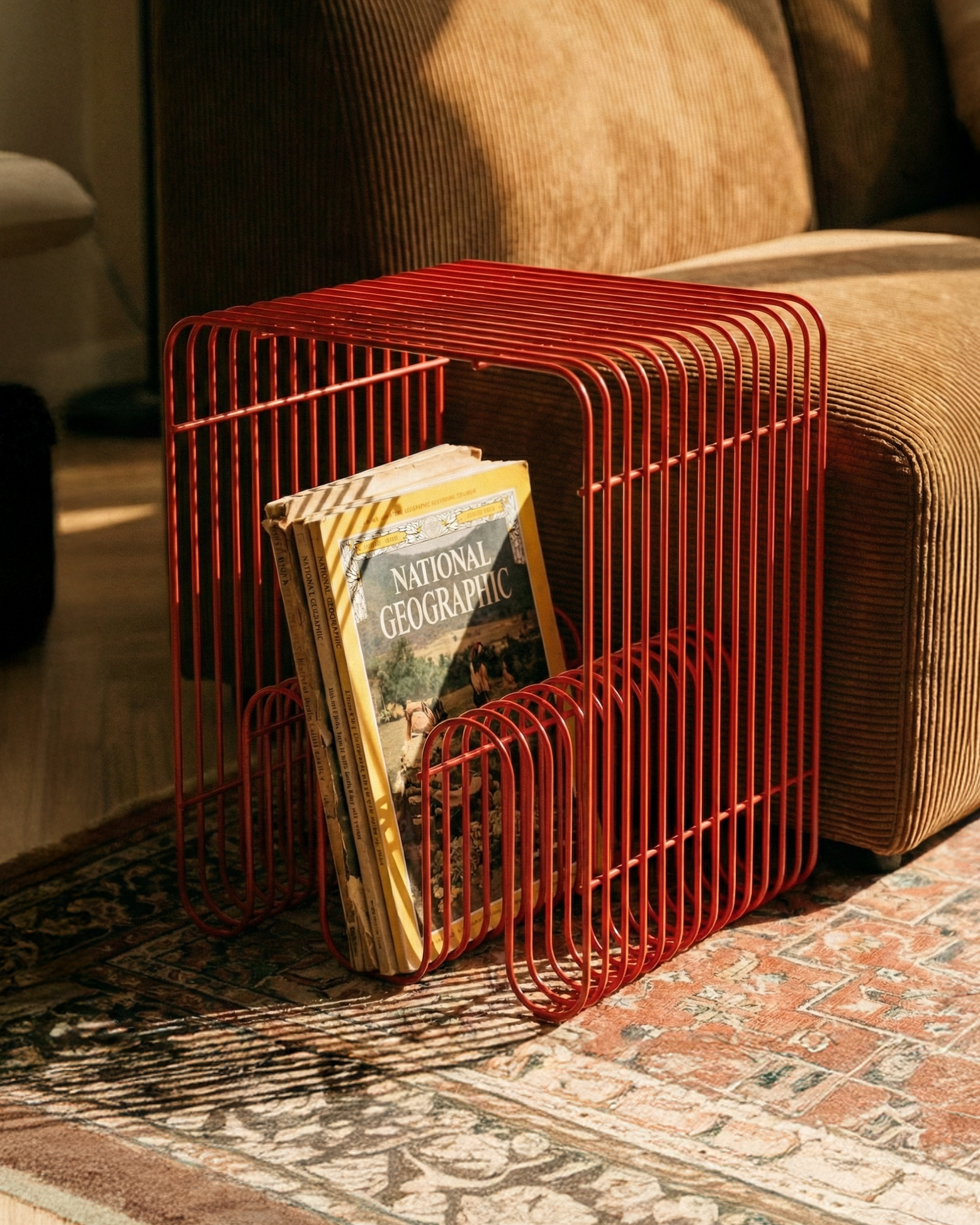 Red wire magazine holder with a book on a patterned rug in a living room.